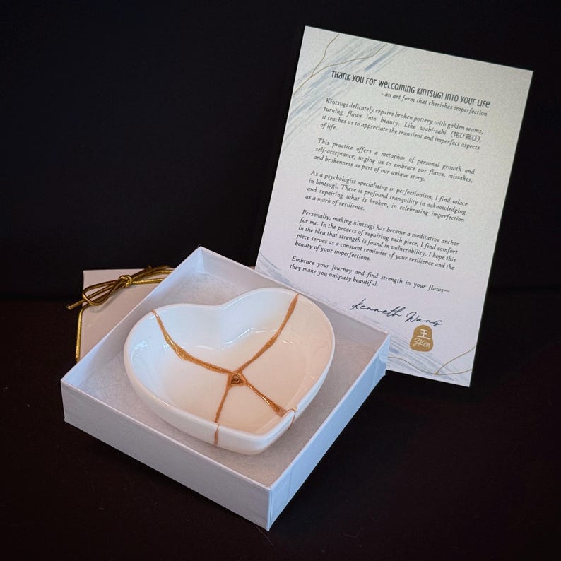 May include: A white heart-shaped ceramic dish featuring gold-coloured kintsugi repair lines, presented in a white gift box with a card titled Thank you for welcoming kintsugi into your life, which explains the art form and includes a signature.