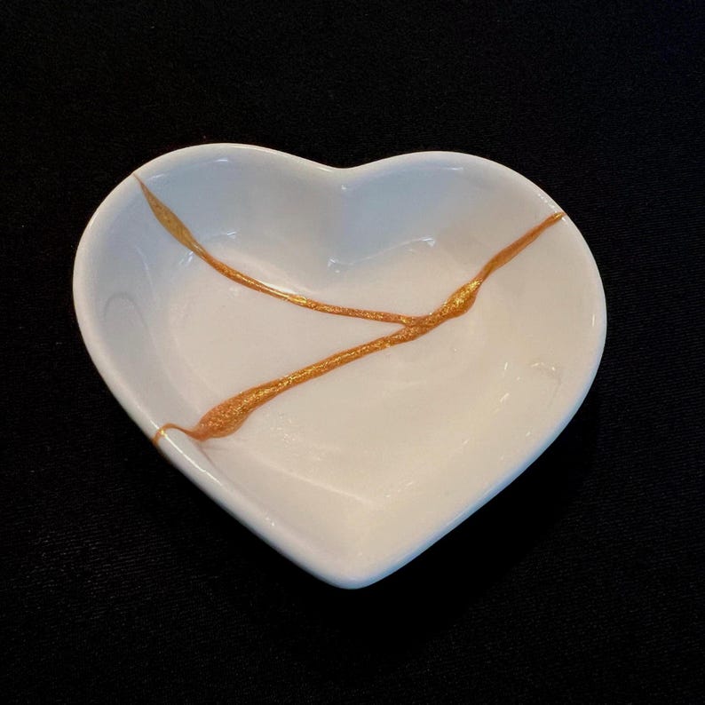 May include: A white heart-shaped ceramic dish featuring a prominent Y-shaped crack repaired with a shimmering gold-coloured adhesive material. The bowl has smooth, curved edges and a glossy finish, highlighting the contrast between the white surface and the gold.