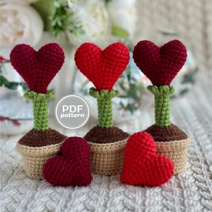 Puede incluir: Tres macetas de flores en forma de corazón de ganchillo con tallos verdes y bases marrones. Hay dos corazones de ganchillo adicionales en primer plano.