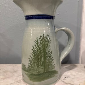 May include: A light blue ceramic pitcher with a glossy finish, featuring a dark blue horizontal band near the rim and a green dendrite-like tree pattern on the body. The pitcher has a curved handle and a flared spout for pouring.