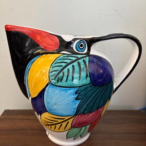 May include: A ceramic pitcher with a bird design. The pitcher features a black beak, a red tip, and a blue eye. The body is decorated with colorful leaf-like shapes in yellow, blue, green, and red. The handle is white.