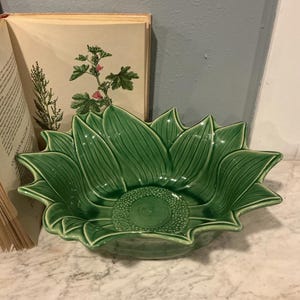 May include: A green ceramic bowl shaped like a sunflower. The bowl has detailed leaf-like edges and a textured center. The bowl is sitting on a marble surface, with a vintage book and botanical illustration in the background.