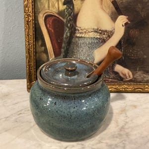 May include: A round, blue-glazed ceramic jar with a matching lid and a small wooden spoon inserted through a notch in the rim. The jar features a speckled finish with darker blue and brown tones, and the lid has a small, centered knob handle.