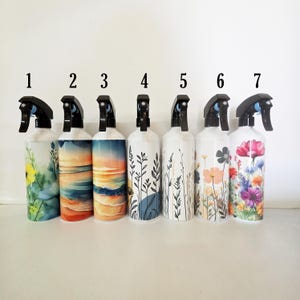 May include: Seven white spray bottles with black triggers, numbered one through seven. Each bottle features a unique design: floral patterns, beach landscapes, and botanical line art. The bottles are arranged in a row against a plain background.