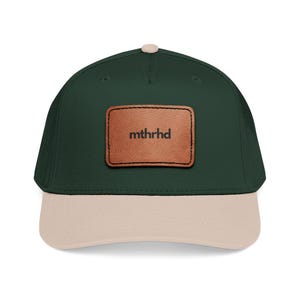 May include: A baseball cap with a dark green crown and a tan brim. A brown leather patch with the word "mthrhd" is centered on the front. The cap has a curved bill and a button on top.