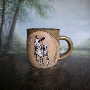 May include: A speckled tan ceramic mug featuring a hand-painted illustration of a young calf standing within a circular frame. The design includes delicate floral accents on the sides of the circle, and the mug has a sturdy, curved handle.