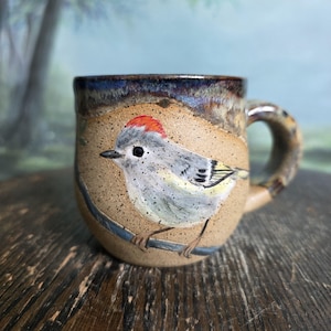 May include: A handmade ceramic mug with a painted bird design. The mug features a tan base color with a blue and brown rim and handle. The bird is gray, white, yellow, and red, perched on a blue branch. The mug is on a wooden surface.