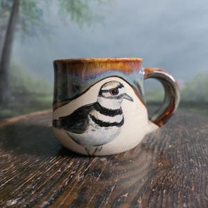 Könnte beinhalten: Handbemalte Keramiktasse mit einer detaillierten Illustration eines Vogels mit schwarz-weißen Markierungen. Die Tasse hat einen braunen und blauen glasierten Rand und Henkel. Ein einzigartiges, handgefertigtes Stück für Kaffee oder Tee.