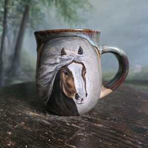 May include: A ceramic mug with a speckled light gray glaze featuring a hand-painted brown and white horse portrait on the front. The mug has a curved handle and a dark brown rim that transitions into a muted green tone at the top edge.