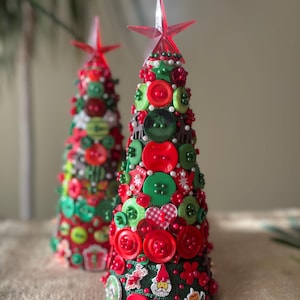 May include: Two decorative Christmas trees. The trees are cone-shaped and covered in red, green, and white buttons, with small decorative stickers. Each tree is topped with a red star. The trees are on a neutral-coloured surface.