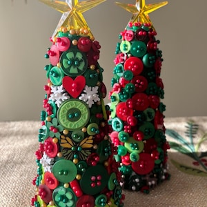 May include: Two handmade Christmas tree decorations. The trees are cone-shaped and covered in red, green, and white buttons. Each tree is topped with a yellow star. The trees are decorated with a heart, snowflakes, and a butterfly.