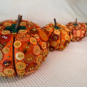 May include: Three decorative pumpkins covered in various orange, brown, and yellow buttons of different sizes and textures. Each pumpkin features a small brown twig stem and is accented with tiny gold beads placed between the buttons.