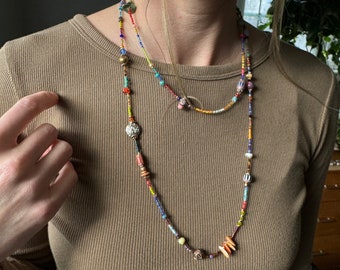 Puede incluir: Un collar de capas con cuentas de colores, incluyendo cuentas pequeñas redondas, cuentas más grandes y algunas cuentas más grandes, planas y rectangulares. El collar está hecho de una variedad de materiales, incluyendo madera, metal y vidrio. El collar lo lleva una persona con el pelo largo y castaño.