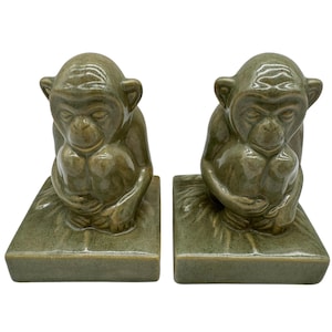 May include: Two olive green ceramic monkey bookends. Each monkey is seated, holding its knees, and resting on a rectangular base. The bookends have a glossy finish and are designed to add a touch of whimsy to any bookshelf.