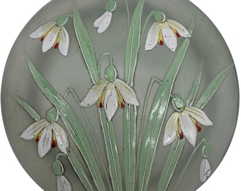 May include: A round, pale green ceramic plate with a raised design of white snowdrop flowers with yellow centers and red accents. The flowers are surrounded by green leaves.