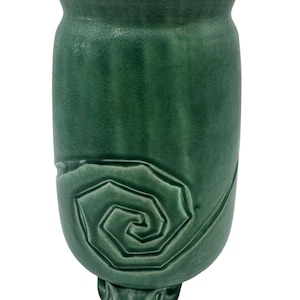 Puede incluir: Un jarrón cilíndrico de cerámica con un esmalte verde bosque mate. El exterior presenta un diseño en espiral en relieve en la sección inferior y un borde acampanado en la parte superior. La base se apoya sobre tres pequeños pies esculpidos.