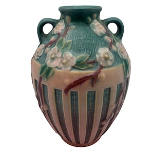 Può includere: Un vaso in ceramica caratterizzato da una sezione superiore color verde acqua con superficie testurizzata e fiori di ciliegio rosa e bianchi in rilievo su rami marroni. Il corpo inferiore presenta strisce verticali in toni alternati di rosa e verde acqua.
