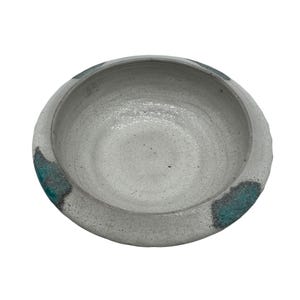 May include: A handmade, round ceramic bowl with a textured, off-white exterior. Teal-colored accents are painted around the rim. The bowl has a wide, shallow design, suitable for serving or display.