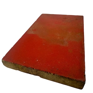 Puede incluir: Un objeto rectangular rojo, ligeramente inclinado. La superficie tiene un aspecto texturizado con variaciones en el color rojo. Los bordes son de un color marrón-amarillento más oscuro.