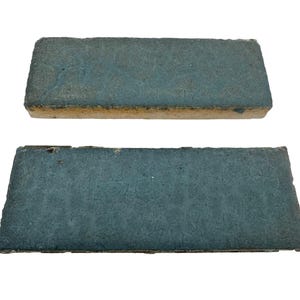 May include: Two rectangular, teal-colored tiles with a textured surface. The tiles have a rough, light brown edge. The tiles are arranged one above the other against a white background. The tiles appear to be made of stone or ceramic.