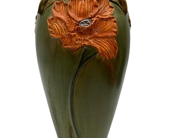 May include: A tall, olive green ceramic vase featuring a textured rim and a prominent, raised relief of a large orange poppy flower with a dark center. The body of the vase displays a smooth, glossy finish with a long, slender stem extending downward.