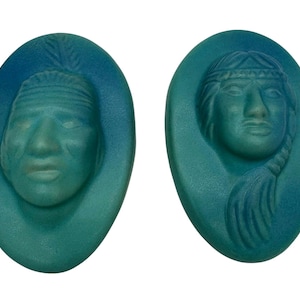 May include: Two oval-shaped teal-colored pendants with relief carvings of Native American faces. One features a headdress, the other a braided hairstyle. The faces are light-colored, contrasting with the teal background.