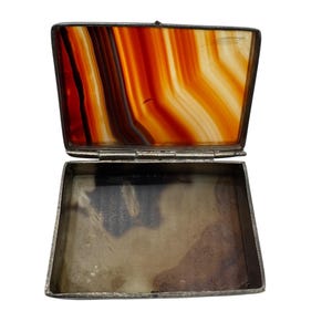 Puede incluir: Caja rectangular abierta de color plateado con tapa abisagrada. La tapa presenta una piedra de ágata pulida con bandas de color naranja, rojo y blanco. El interior de la caja está vacío y tiene un aspecto deslustrado.