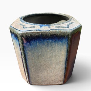 May include: A hexagonal ceramic planter featuring a reactive glaze with a blend of deep blue, cream, and earthy brown tones. The rim displays a textured, light-colored surface with subtle geometric patterns, while the body transitions into smooth, glossy blue and brown gradients.