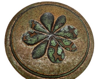 May include: A round, ceramic trivet with a raised floral design. The trivet features a central flower with six green petals and a brown center. The background is a mottled brown and green, with a dark green border. This decorative piece is suitable for kitchen use.