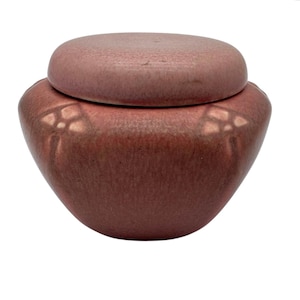 May include: A dusty rose ceramic jar with a matching circular lid. The vessel features a rounded, bulbous body with subtle, embossed floral motifs on the sides. The glaze has a matte, textured appearance with slight variations in color tone.