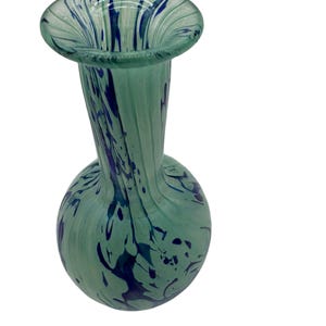 Puede incluir: Un jarrón de vidrio verde con vetas azules y blancas. El jarrón tiene una base redonda y un cuello estrecho.