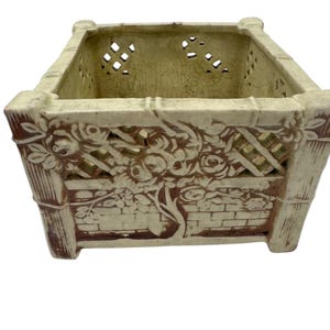 Puede incluir: Un macetero cuadrado de cerámica con un esmalte marrón y blanco. El macetero tiene un diseño de celosía y un motivo floral. El macetero está diseñado para parecer un enrejado con rosas que crecen en él.