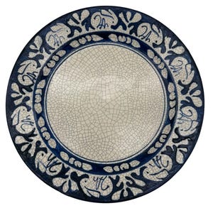 May include: A round ceramic plate featuring a central cream-colored area with a fine crackle glaze finish. The wide border is decorated with a repeating pattern of dark blue stylized rabbits and abstract floral shapes against a cream background.