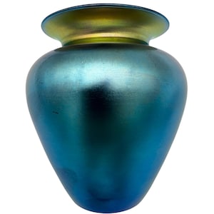 Puede incluir: Un jarrón de vidrio azul iridiscente con un cuerpo ancho y redondeado y un borde acampanado. El jarrón tiene un brillo metálico, con un degradado de colores de verde azulado a dorado en la parte superior. Es un artículo decorativo.