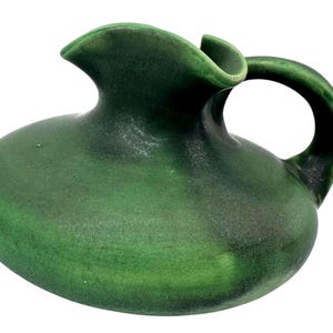 May include: A small, green ceramic pitcher with a rounded body and a curved handle. The pitcher has a spout for pouring and a textured surface. The dark green color gives it a rustic, vintage feel. It is likely used for serving liquids.