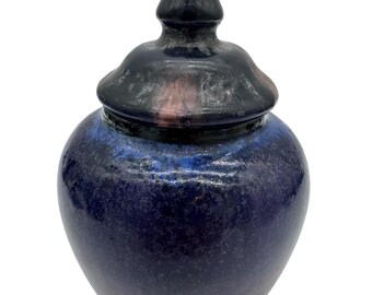 May include: A ceramic jar with a removable lid featuring a pointed finial. The vessel has a rounded body with a glossy glaze in deep shades of navy blue and black, transitioning to subtle hints of reddish-pink near the base and lid edges.
