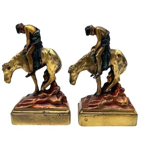 May include: A pair of antique bronze bookends. Each bookend features a figure on horseback, with the rider draped in a green cloth. The horse and rider are gold-toned, mounted on a reddish-brown base.