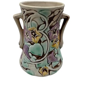 Pode incluir: Um vaso de cerâmica com base creme e duas alças. Apresenta um design floral em tons de verde, roxo e amarelo. O design é colocado sobre um fundo de redemoinhos castanhos e creme. Um item decorativo.