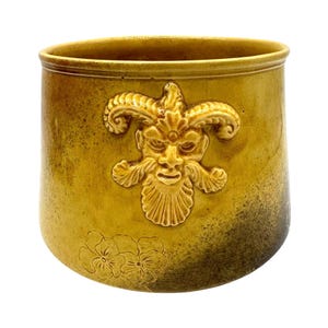 May include: A cylindrical ceramic pot with a mustard yellow glaze and a dark brown gradient at the base. The exterior features a raised relief of a horned, bearded face centered on the front, with a small floral pattern embossed near the bottom edge.
