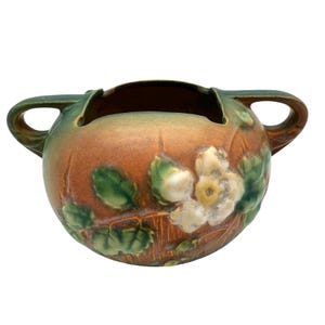 May include: A ceramic bowl with two handles, featuring a floral design. The bowl has a brown base with green and white accents. The handles and rim are green. The floral design includes white flowers and green leaves.