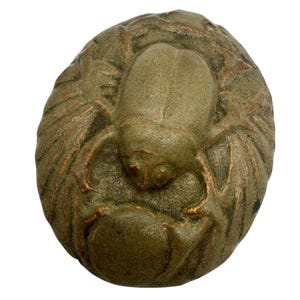 May include: A detailed, olive-green sculpture of a scarab beetle, a symbol of ancient Egypt. The beetle is centered on a textured, oval base with intricate wing details. The sculpture has a weathered, aged appearance.