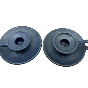 May include: Two dark blue ceramic candle holders with a matte, textured finish. Each holder features a circular base, a central cup for a candle, and a small, curved handle extending from the side, designed for easy carrying and placement.