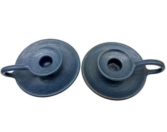 May include: Two dark blue ceramic candle holders with a matte, textured finish. Each holder features a circular base, a central cup for a candle, and a small, curved handle extending from the side, designed for easy carrying and placement.