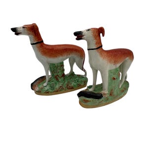 Puede incluir: Dos figuras de perros galgos de cerámica. Los perros tienen cuerpos de color marrón rojizo, patas blancas y collares y narices negros. Se paran sobre bases verdes y marrones. Las figuras están sobre un fondo blanco.