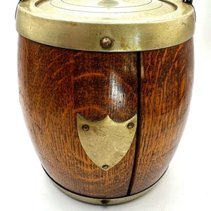 May include: A wooden barrel-shaped biscuit tin with a brass lid and a brass shield on the front. The lid has a small brass knob on top.