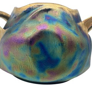 May include: A ceramic vase with two handles, featuring a unique iridescent glaze in shades of blue, green, and purple. The vase has a textured surface and a gold rim.