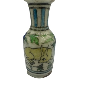 Op de afbeelding: Een kleine, antieke keramische vaas met een crèmekleurige basis. De vaas heeft een gele dierenillustratie, blauwe en groene bloemaccenten en blauwe verticale strepen aan de bovenkant. De vaas is ongeveer 13 cm hoog.