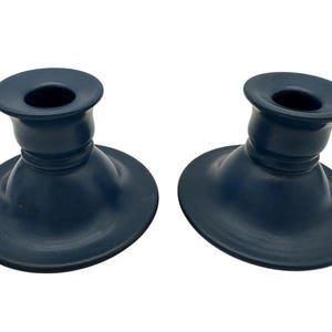 May include: A pair of dark navy blue ceramic candle holders with a smooth matte finish. Each holder features a wide, circular base that tapers upward into a cylindrical neck with a flared rim designed to securely hold a standard taper candle.
