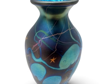 May include: An iridescent glass vase featuring a dark blue and black base with swirling light blue organic shapes and thin, delicate pink lines. The vase has a flared rim and a smooth, reflective surface with small orange star-like accents.