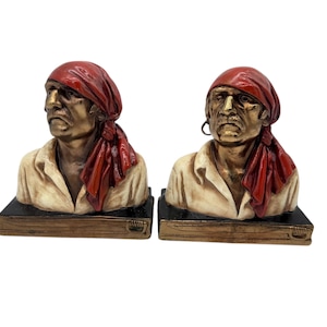 Puede incluir: Dos bustos de bronce que representan a hombres con pañuelos rojos y camisas crema. Los bustos están montados sobre bases rectangulares negras con ribetes dorados, con un aspecto vintage.